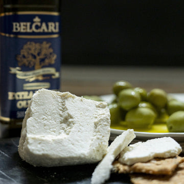 Cuts of Ricotta cheese on biscuits with green olives and a bottle of olive oil in the background.