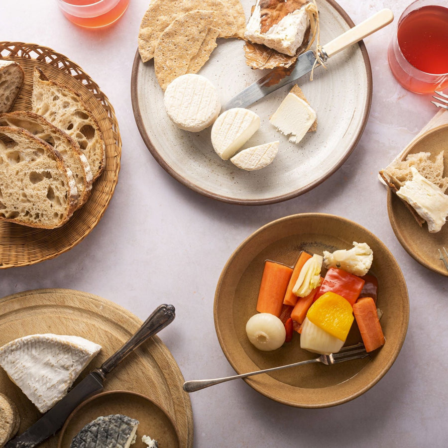 A table laid out with plates of cheese and pickles, a basket of bread and a board of several other cheeses