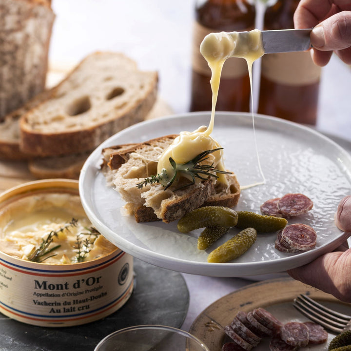 Soft, melting Vacherin cheese running off a knife onto a slice of bread on plate, with some cornichons and slices of salami.